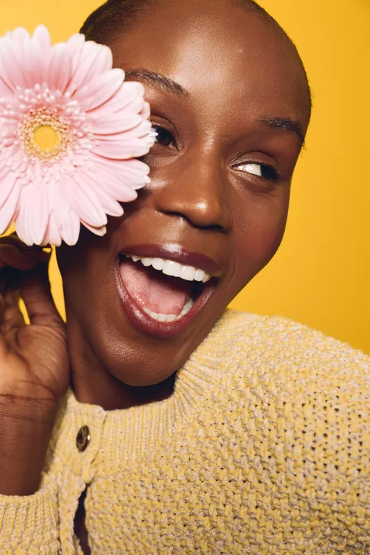 Happy African woman with dark skin holds pink gerbera flower near face, smiling with open mouth. Bright yellow background and cozy knitted sweater create warm positive mood.