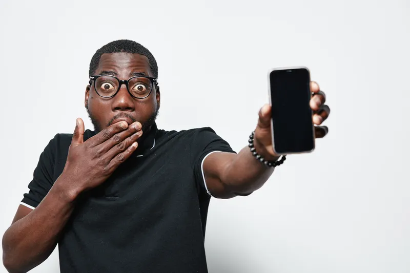 Black man with dark skin and glasses showing smartphone with blank screen and surprised expression on face in studio with white background.