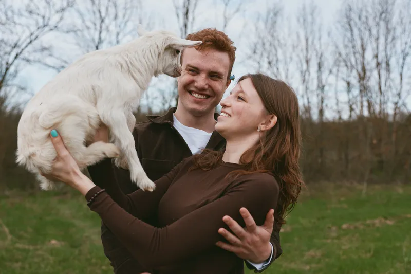 A cheerful couple outdoors holds a white goat kid, smiling as they lift the playful animal. Warm, candid lifestyle moment showing bonding, affection, and gentle animal companionship in a park.