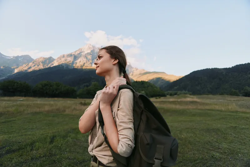 Bright afternoon outdoors, a woman with a backpack stands in a grassy field, mountains in the distance, calm expression, soft breeze, natural tones, lifestyle moment, nature theme