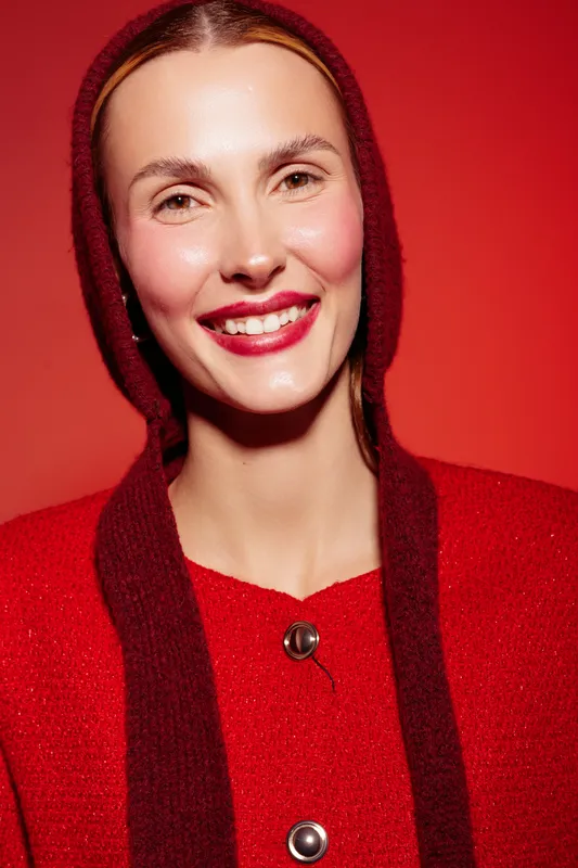 Smiling woman with red lips and natural makeup wears red sweater and hood against red background. Concept of fashion, beauty, and confident female portrait with warm studio lighting.
