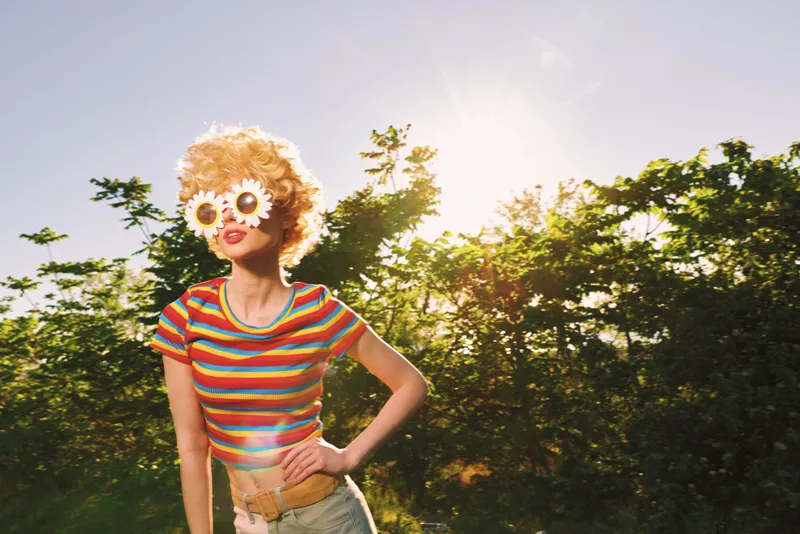 Young woman with curly hair wears oversized flower sunglasses and a colorful striped top in a sunlit garden, radiating playful retro charm and a carefree summer mood.