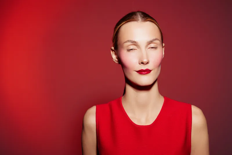 Young woman with closed eyes wearing red dress and red lipstick stands against red background. Concept of fashion, beauty, makeup, and monochrome style portrait.