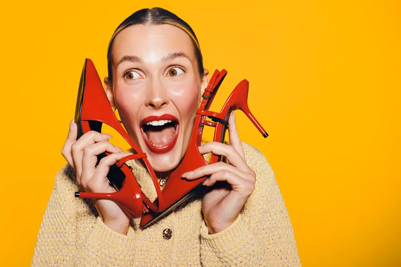 Excited woman holds red high heel shoes near face, smiling widely on bright yellow background. Concept of fashion, footwear, style, and joyful emotions in studio lighting.