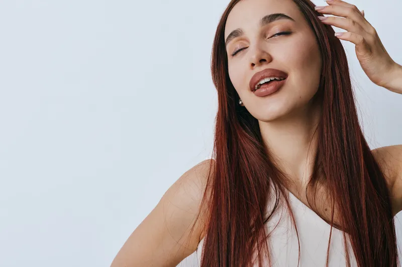 Woman with long red hair and closed eyes posing with hand near head on light background. Beauty, fashion and portrait of female model with natural makeup and relaxed expression.