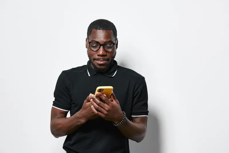 African man with dark skin and glasses using smartphone in black polo shirt on white background. Casual technology, communication and digital lifestyle concept with focused expression.