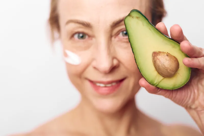 Smiling mature woman holds fresh avocado half near face with natural cream on cheek. Concept of organic skincare, healthy lifestyle, anti aging and natural beauty care.