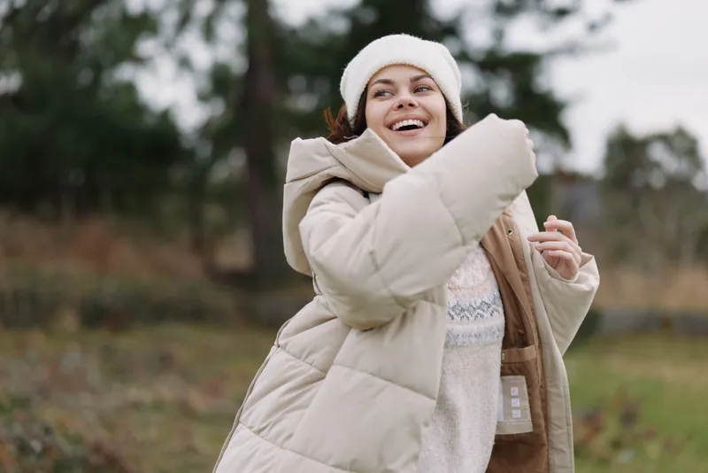 Woman wearing a light beige winter coat smiles joyfully outdoors, enjoying a breezy day in a field as a candid moment of warmth and comfort in chilly weather