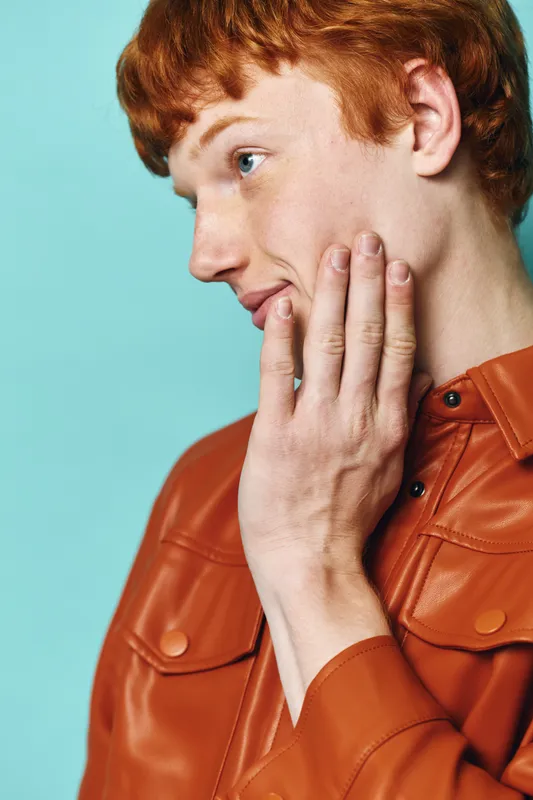 Young man with red hair wearing a brown leather jacket touching his cheek with hand looking thoughtful on blue background, closeup side portrait with casual style.