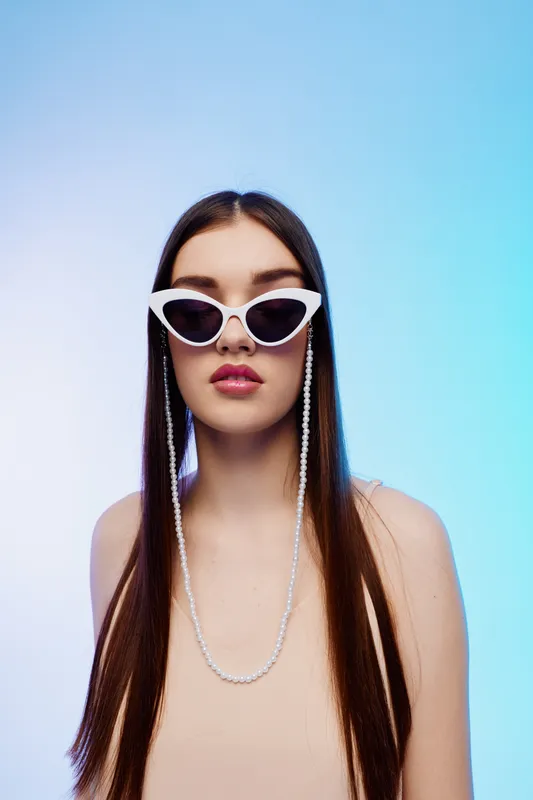 Young woman with long straight hair wearing white cat eye sunglasses and a pearl necklace poses against a gradient blue background. Concept of beauty fashion style.