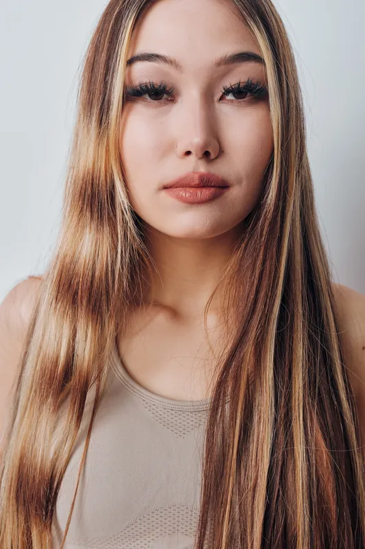 Portrait of young woman with long straight hair and natural makeup in neutral top against light background, looking at camera with calm expression and relaxed posture.