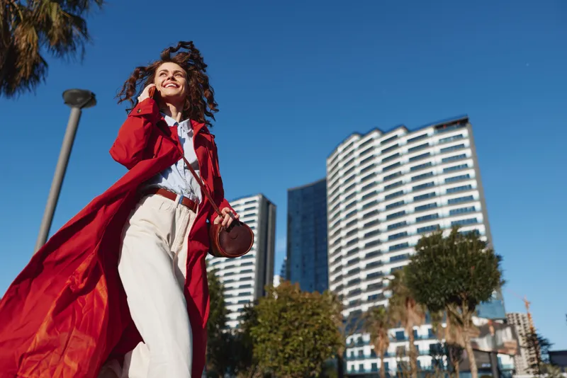 Cheerful woman outdoors wearing a red coat, walking under a bright blue sky, with modern city towers in the background, catching a warm sunlit moment and a carefree smile today