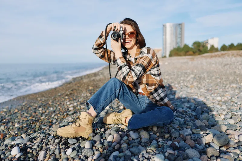 Female photographer on a pebble beach capturing a candid moment with a compact camera, wearing a plaid shirt and jeans, enjoying sunny weather and the sea breeze.