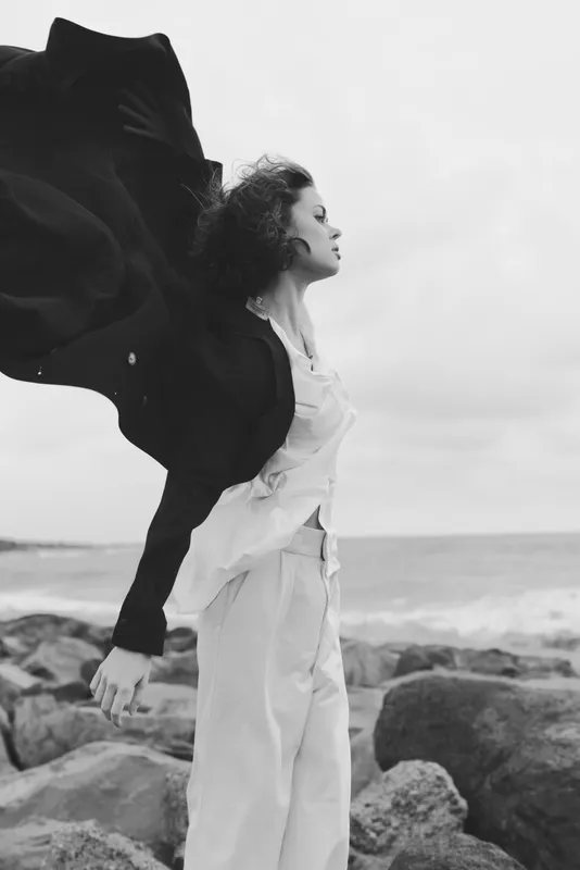 Wind swept, white outfit flares against a rocky shoreline in monochrome, a dramatic sea backdrop and flowing fabric capturing movement and fashion on the coast