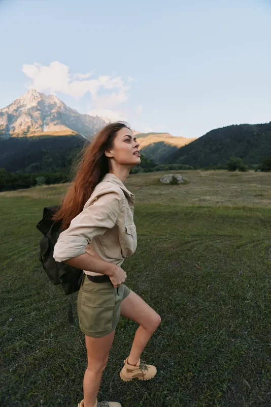 Young woman with long hair stands on a grassy field looking at a mountain during sunset, enjoying nature and outdoor adventure with a backpack.