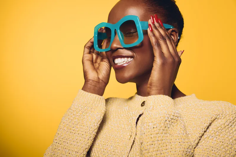 Happy African woman with dark skin wears oversized blue glasses and beige sweater smiling with closed eyes on yellow background. Concept of fashion, beauty, and positive mood.