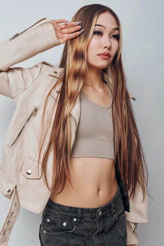 Young woman with long straight hair wearing light jacket and crop top posing with hand near head on gray background with confident expression and casual style.