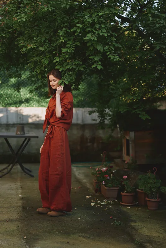 One woman in a rust red dress stands outdoors in a lush garden, talking on her phone with a calm, relaxed expression under leafy shade
