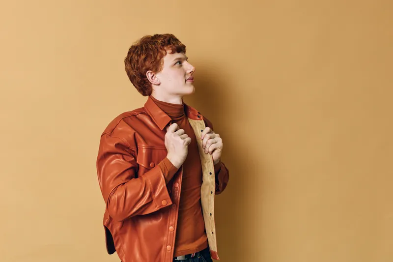 Young man with red hair wearing brown leather jacket and turtleneck looking sideways thoughtful with beige background fashion style casual portrait profile mood.
