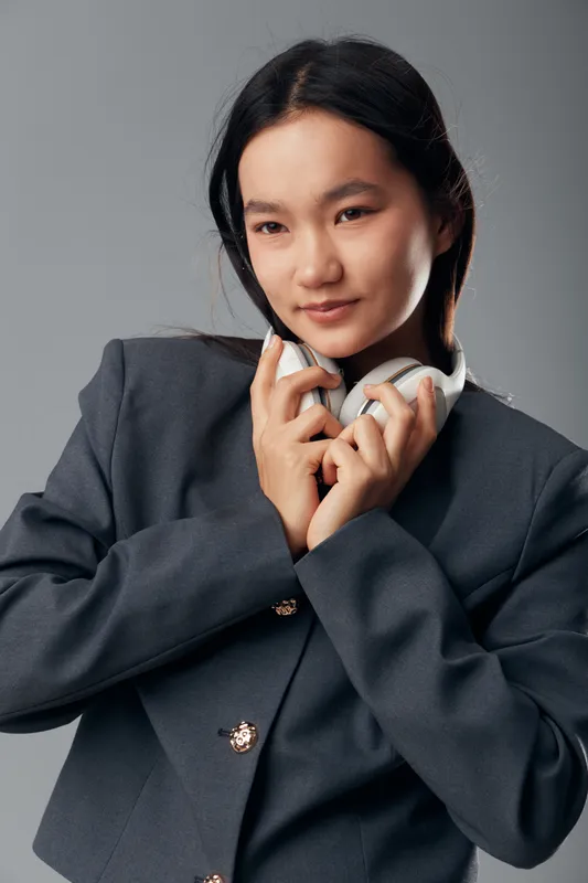 Young woman with headphones wearing dark blazer looking down thoughtfully isolated on grey background. Portrait of casual female adult with long hair in studio with minimal expression.
