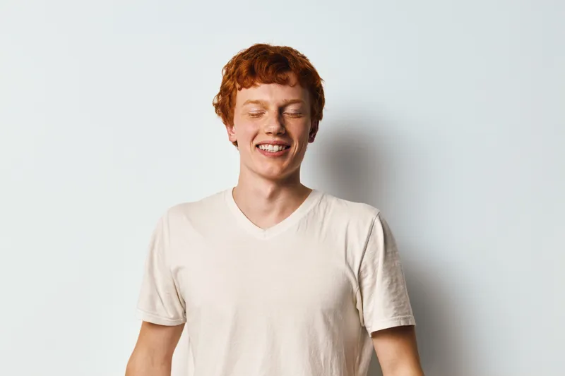 Young man with red hair and closed eyes wearing white t shirt smiling in front of white background. Happy teenager or adult male with freckles and casual style portrait in studio indoors.