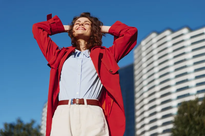 Cheerful woman outdoors, wearing a red jacket over a white shirt, blue sky backdrop, confident pose, sunlit scene, urban backdrop, bright colors, natural light, energetic mood, carefree attitude