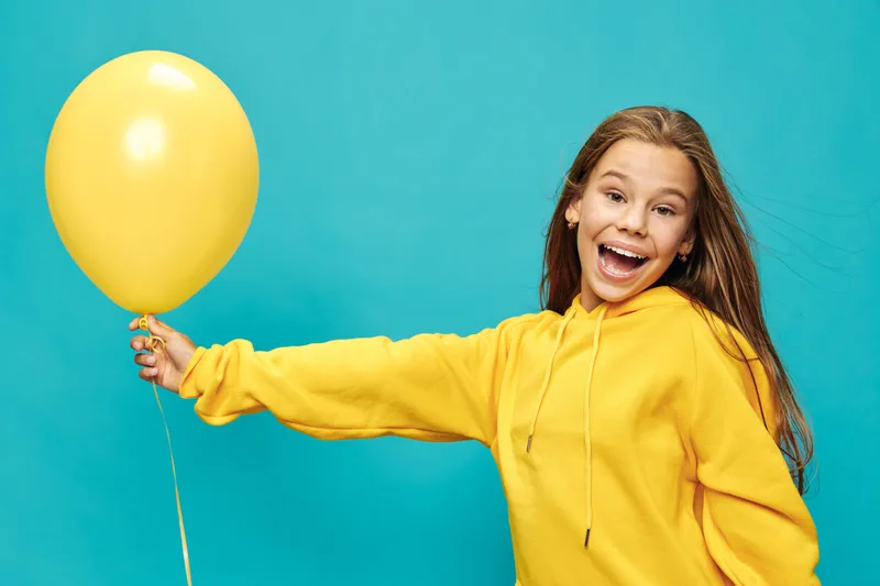 Girl in yellow hoodie holding yellow balloon with excited smile on blue background. Teenage child with long hair posing cheerful for celebration, joy and fun with colorful party prop.