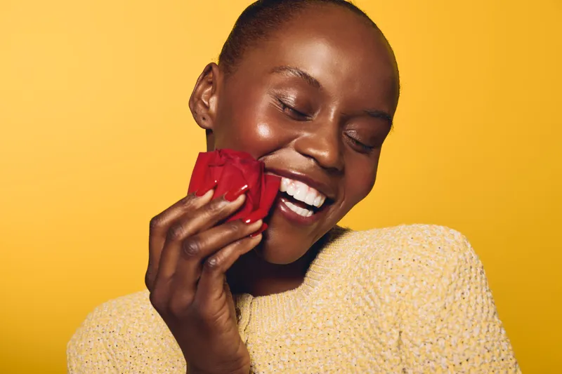 Happy African woman with dark skin smiling and holding a red rose near her face. Close-up on yellow background with warm lighting. Concept of joy, beauty, and natural skincare.