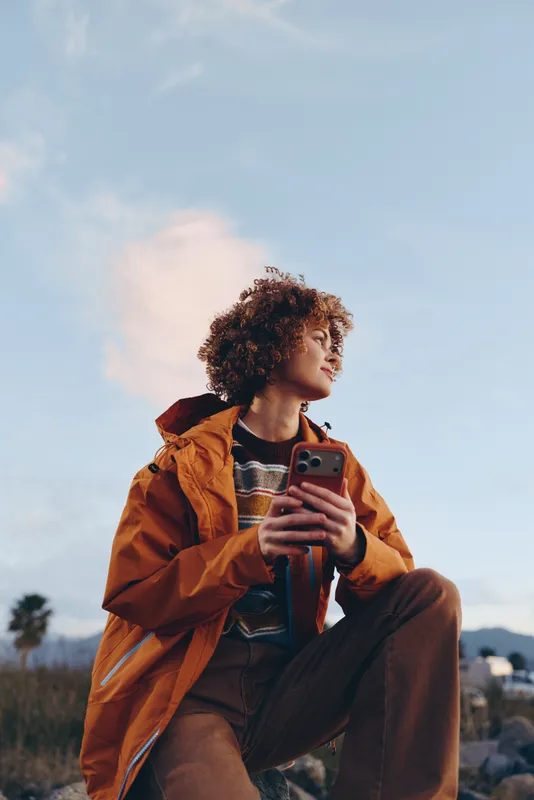 Woman outdoors, curly hair, orange jacket, holding smartphone while sitting on street steps, relaxed lifestyle in daylight sky, soft breeze, candid portrait, warm colors natural tones