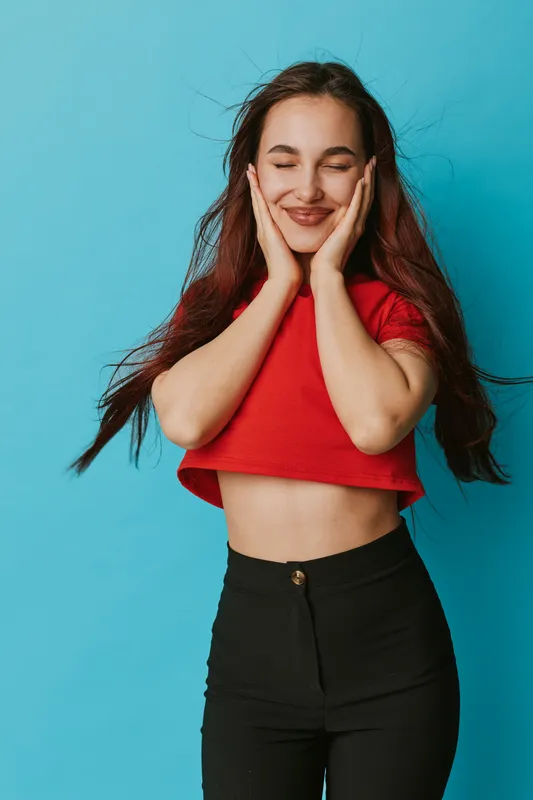 Young woman with long hair wearing red crop top and black pants posing with eyes closed and hands on cheeks against blue background, smiling and enjoying moment.