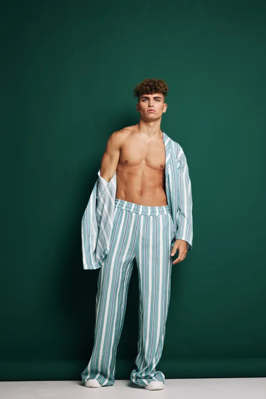 Young man with curly hair wearing striped pajamas standing against green background with shirt open showing bare chest and relaxed pose in studio setting.