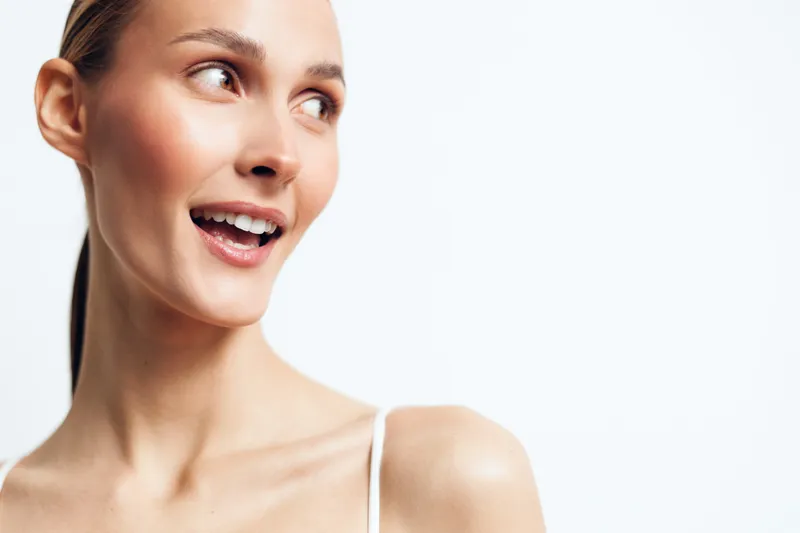 Smiling woman looking sideways with joyful expression, clear skin, natural makeup, wearing white strap top, bright studio background, positive mood, healthy lifestyle concept