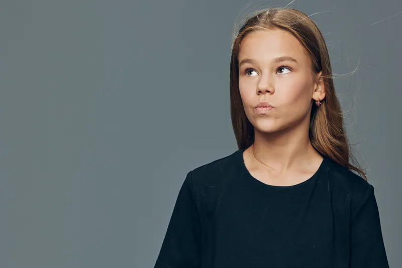 Girl teenager looking aside with thoughtful expression in studio, isolated on grey background. Portrait of young female with long hair wearing dark shirt, pensive mood and contemplation.