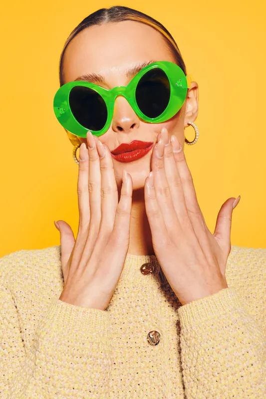 Young woman with bright red lipstick wearing large green sunglasses and yellow sweater poses with hands near face on yellow background. Concept of fashion, style, and summer accessories.