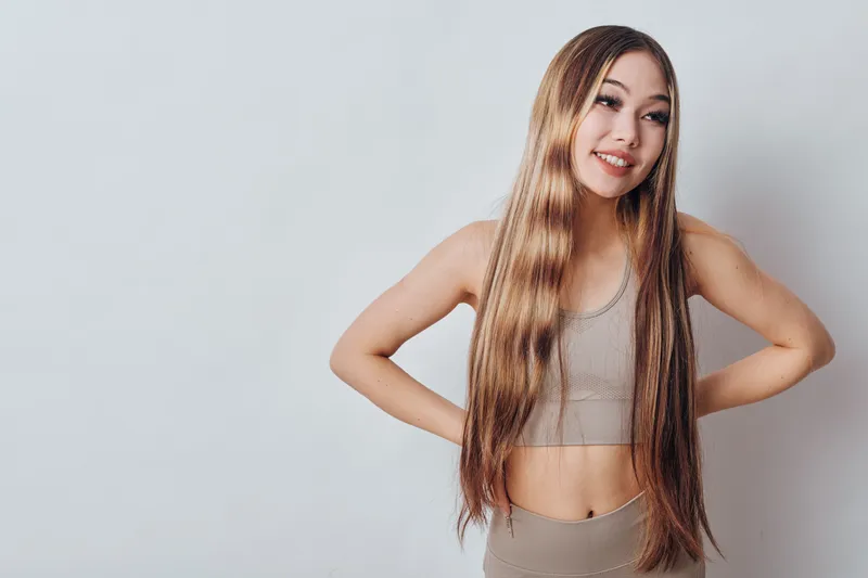 Young woman with long straight hair and casual beige outfit poses smiling with hands on hips against plain light background in studio photography portrait.