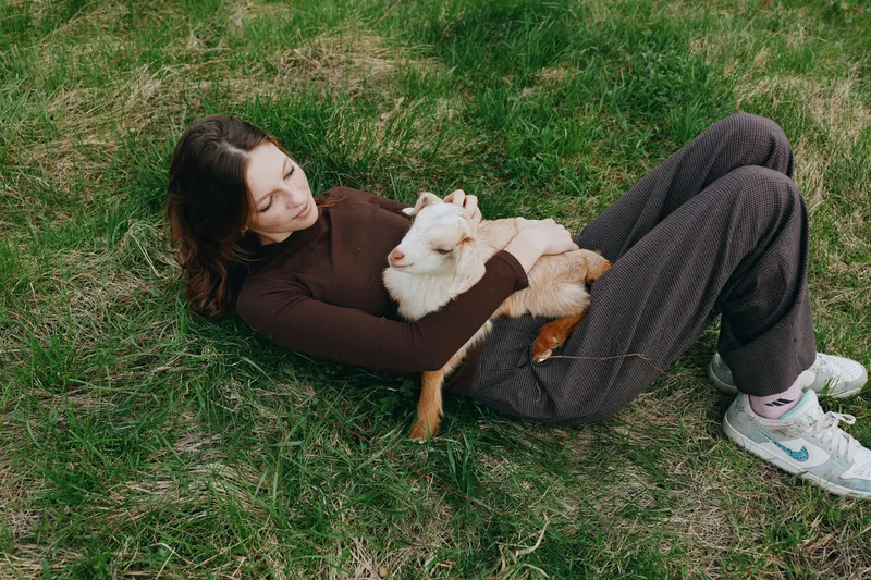 A woman lies on the grass wearing a brown sweater and loose pants, cuddling a fluffy dog. The scene conveys relaxed outdoor lifestyle, affection, and simple moments of everyday happiness.
