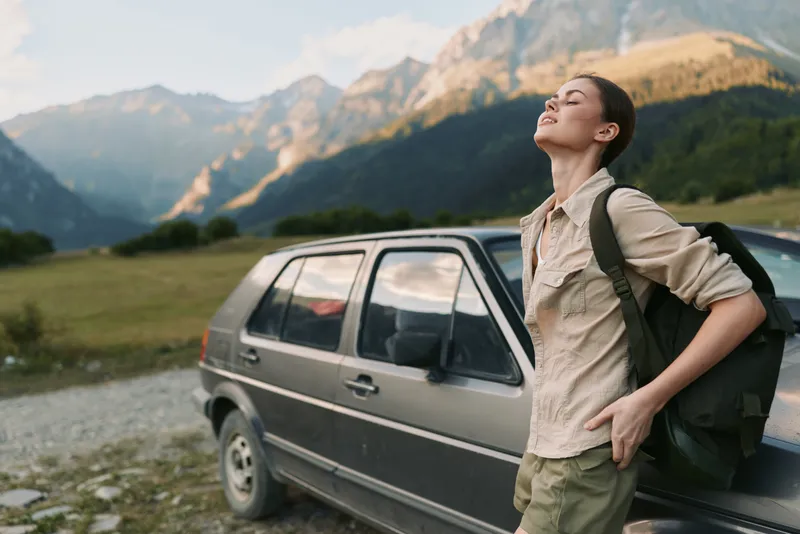 A young woman with a backpack stands beside a weathered car in a rugged mountain valley, soaking in fresh air, wide skies, and vast scenic beauty.
