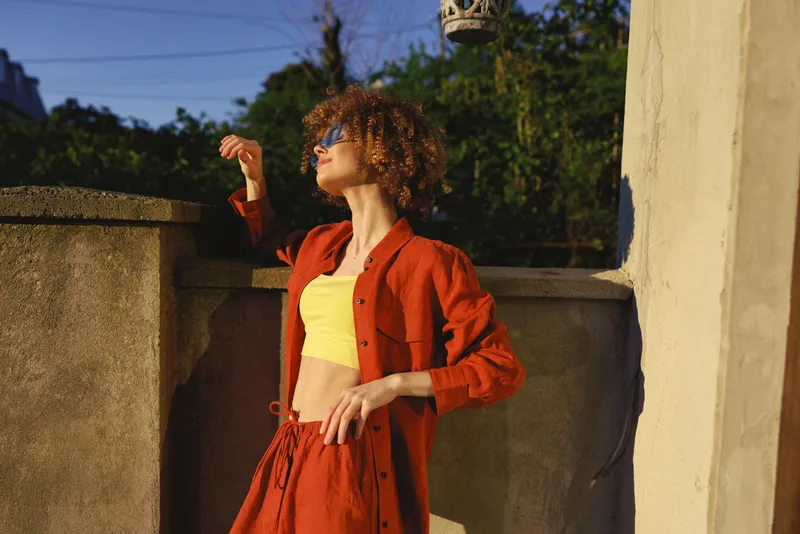 Young woman with curly hair poses outdoors in a bright red outfit and yellow top, enjoying sunlight and relaxed breeze. She leans against a stone wall, sunglasses on, expressing confidence