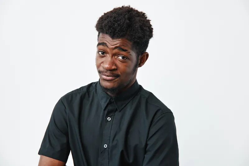 Young black man with dark skin wearing black shirt posing with skeptical expression on white background. African ethnicity, casual style and confident attitude in studio portrait.