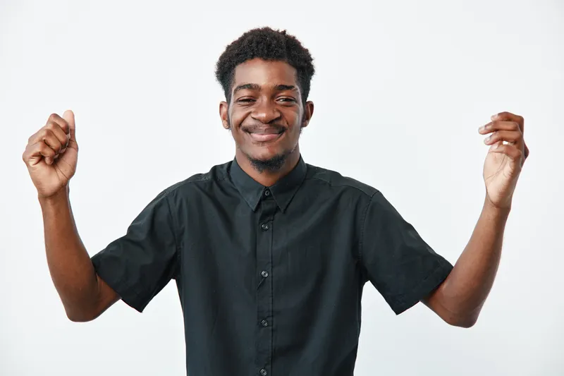 Young black man with dark skin wearing black shirt smiling with closed eyes and raised hands isolated on white background. Positive emotion and casual style portrait.