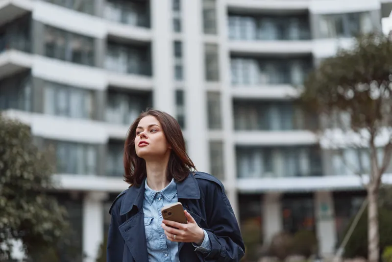 Urban street portrait of a young brunette woman in a denim jacket holding a smartphone, standing in an outdoor city setting with a thoughtful, confident expression.