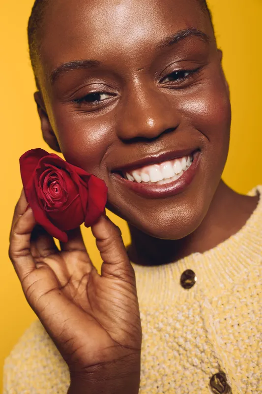 Smiling black woman with dark skin holds red rose near face, wearing yellow sweater. Close-up portrait on yellow background. Concept for beauty, happiness, and natural skincare.