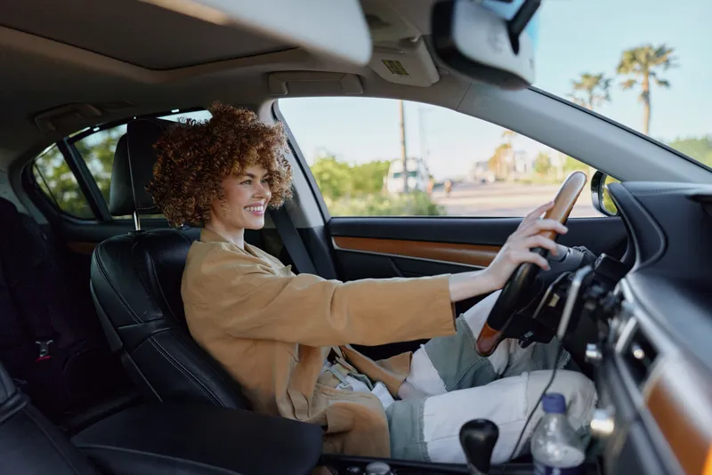 Young woman with curly hair drives a car, smiling softly as she concentrates on the road, wearing a beige jacket inside a sunlit cabin, conveying casual independence and everyday mobility with a