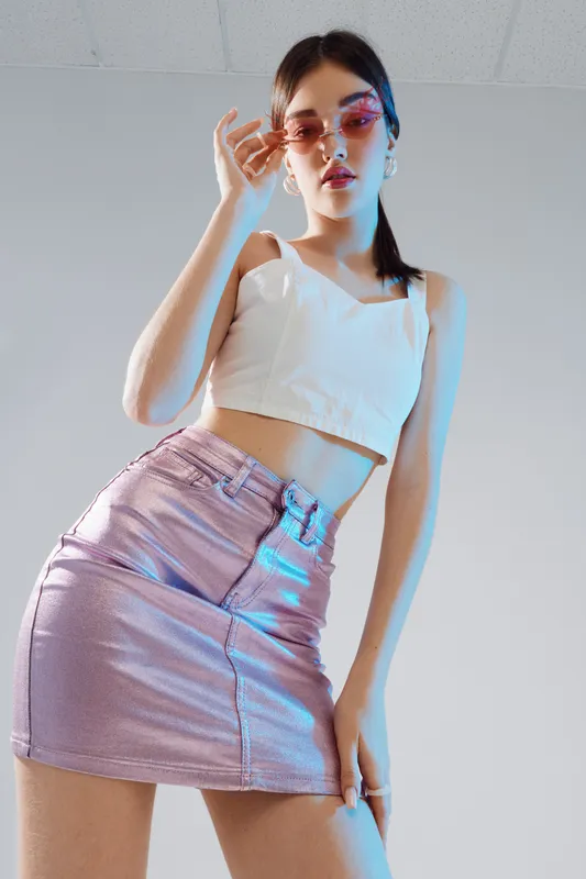 Young woman in a shiny pink skirt and white crop top poses confidently wearing pink sunglasses. Studio shot with soft lighting on a neutral background. Concept of fashion beauty style.