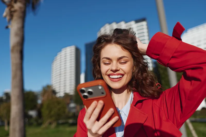 Young woman in a red blazer smiles while taking a selfie with a smartphone against a sunny cityscape, tall buildings behind her, energetic mood and confident pose.