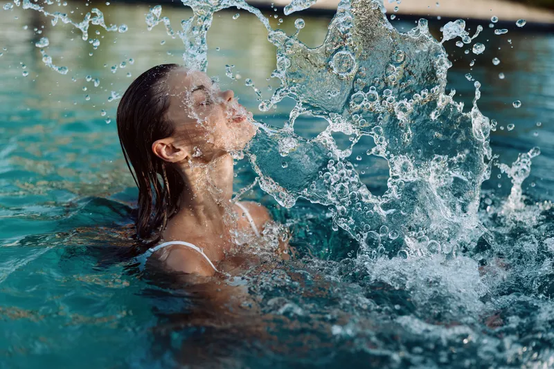 Woman in a pool splashes water, smiling softly. Outdoor bright scene, refreshing moment of wellness and leisure.