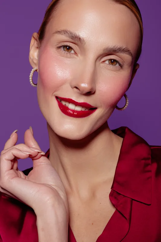 Close-up portrait of a smiling woman with red lipstick and hoop earrings wearing a red blouse on purple background. Concept of beauty, makeup, fashion, and confident expression.
