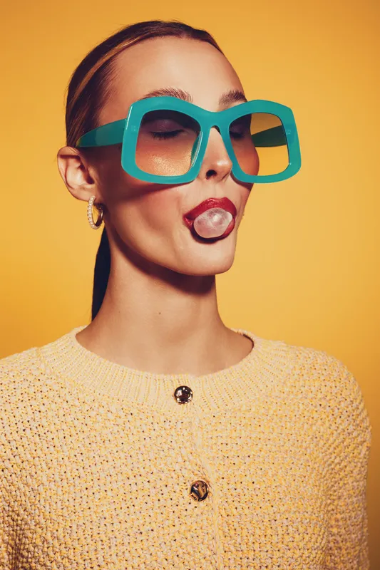 Young woman with slicked hair wearing large turquoise sunglasses blows a bubblegum bubble. She wears a yellow knitted sweater against a bright yellow background. Fun and retro style.