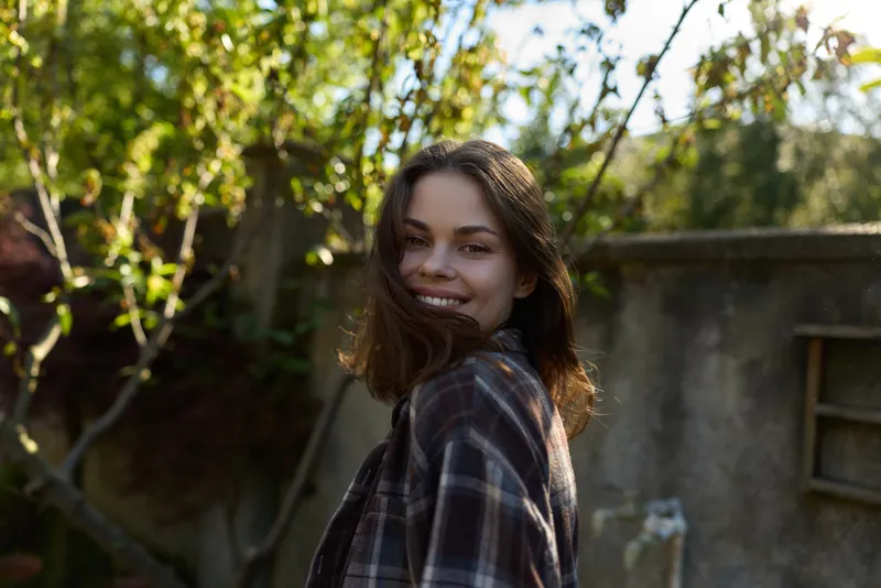 Cheerful woman outdoors, turning back and smiling, sunlit garden setting, casual plaid shirt, relaxed lifestyle moment, natural colors, soft breeze, candid portrait