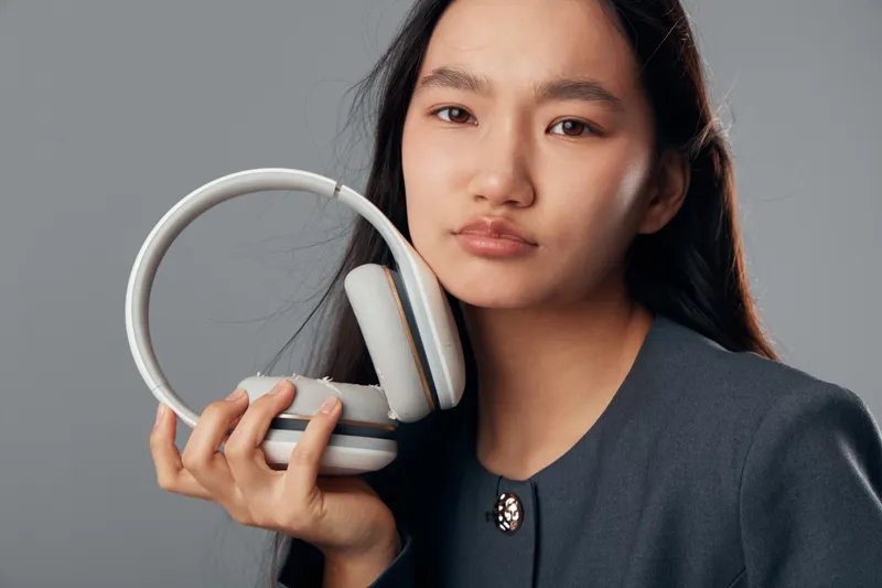 Young woman holding gray headphones with long dark hair and neutral expression in studio. Fashionable female model wearing dark clothes on gray background with technology accessory for music.